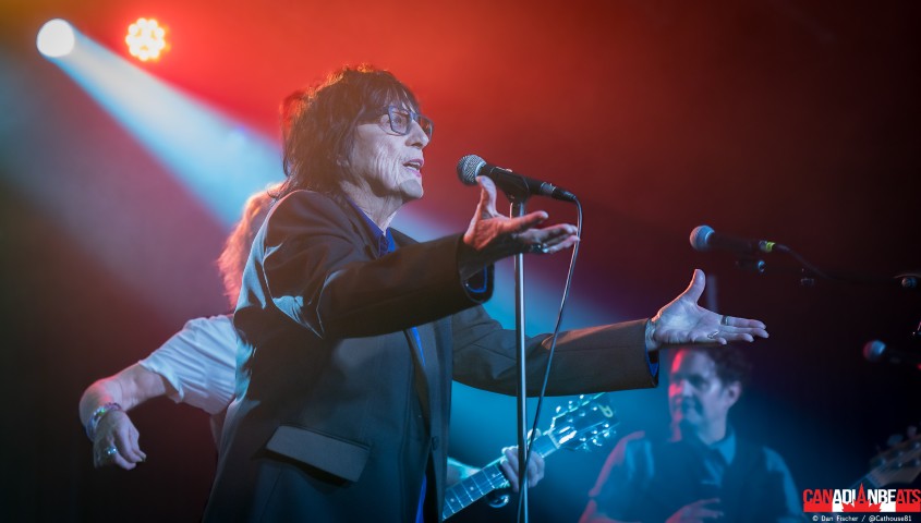 Carole Pope of Rough Trade singing into a microphone with her arms extended under red and blue stage lights in Waterloo.